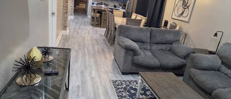 A modern living room with a grey sofa, a glass coffee table, and a patterned rug. A kitchen with dark cabinets and a microwave is visible in the background.