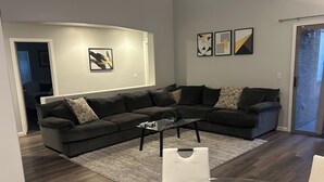 A modern living room with a sectional sofa, a glass coffee table, and a dining table with chairs. There are abstract wall art pieces and a door leading to another room. - Avondale Retreat with Pool near Stadium, Top Golf and Spring Training (Avondale)