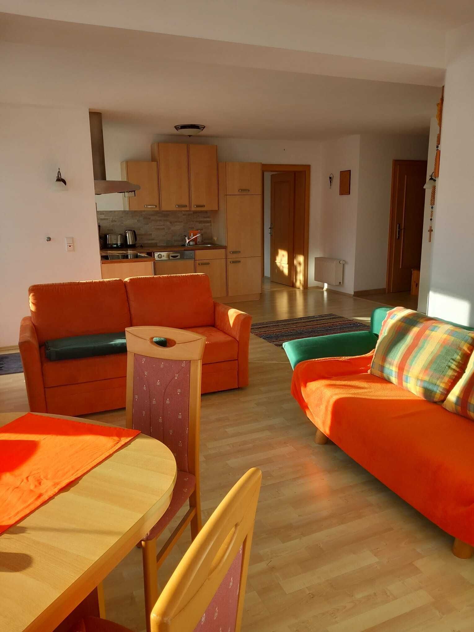 Una sala de estar moderna con sofás naranja, una mesa de comedor de madera y una cocina al fondo.