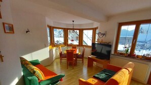 A spacious living room with a wooden floor, a green and orange sofa, a television, and large windows offering a view of a snowy landscape. - House at the edge of the forest Thiersee (Thiersee)