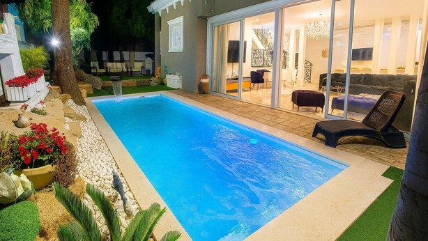 Une piscine avec un espace patio, un coin salon couvert et une vue sur un intérieur bien éclairé.