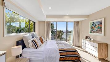 A bedroom with a large bed, a nightstand, a dresser, and a window with a view of greenery and buildings.