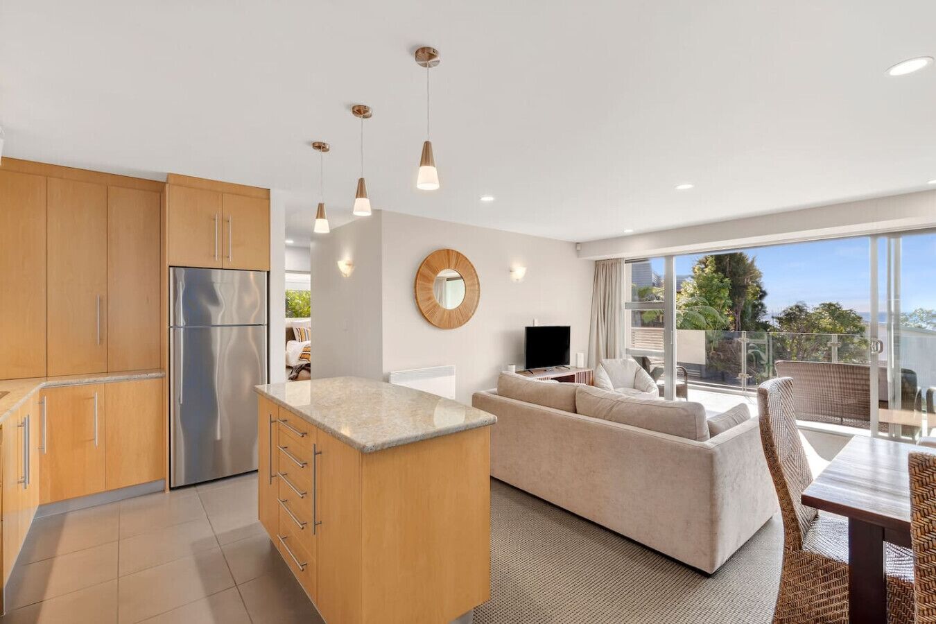 A modern kitchen with wooden cabinets, a central island, and pendant lighting. There is a living area with a sofa and a TV, and a balcony with a view.
