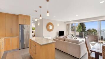 A modern kitchen with wooden cabinets, a central island, and pendant lighting. There is a living area with a sofa and a TV, and a balcony with a view.