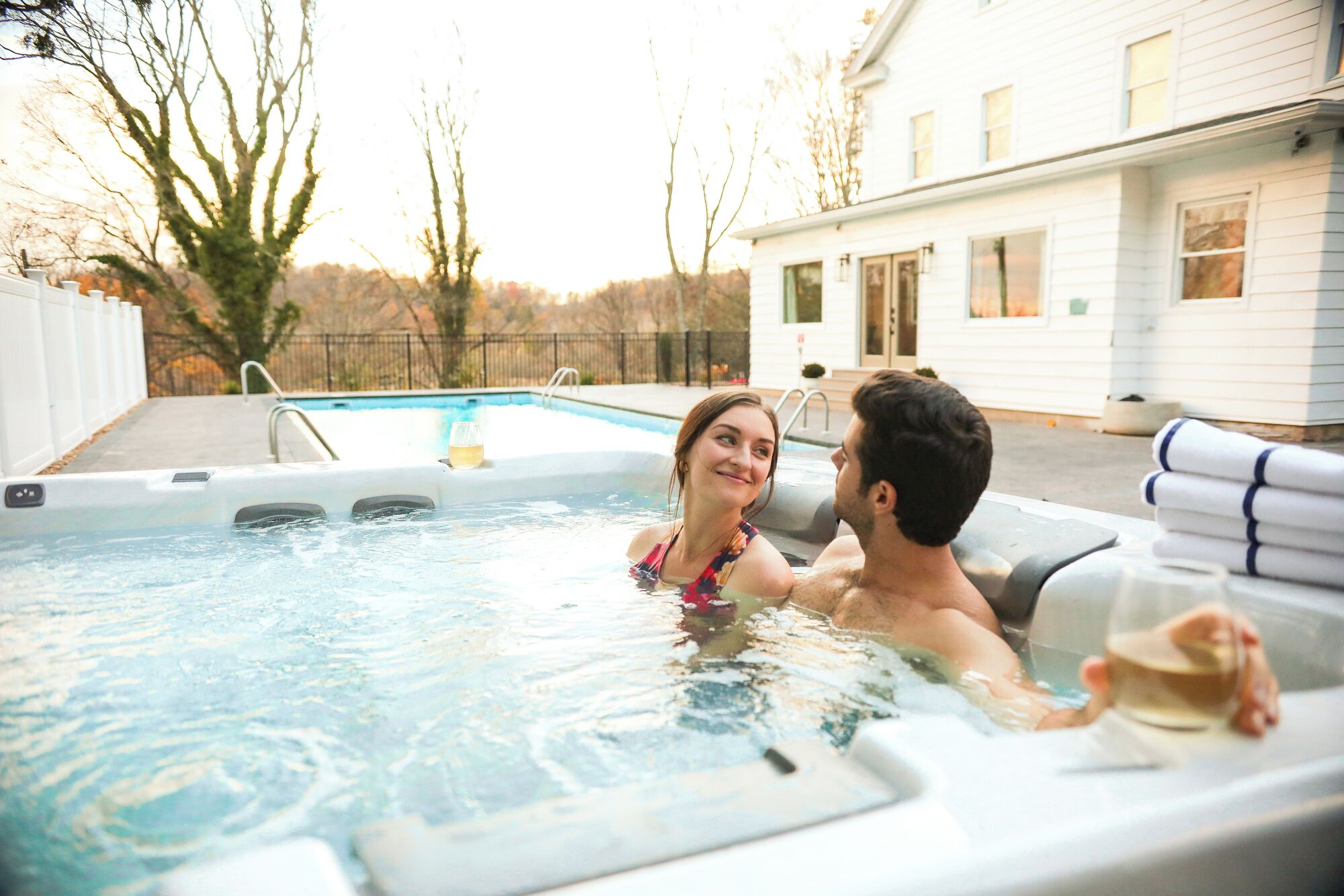 A hot tub with a pool in the background, a house, and trees.
