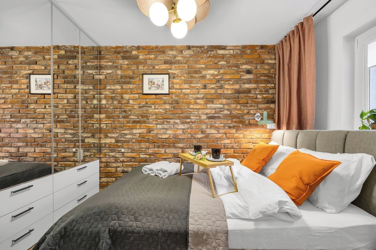A modern bedroom with a brick wall, a bed with white and orange pillows, a bedside table with a tray, and a mirror.