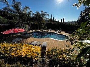 Una piscina circondata da poltrone prendisole e un ombrellone rosso, immersa nel verde e alberi alti.