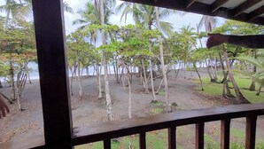 Une vue depuis un espace abrité, doté d’une balustrade en bois, donnant sur une plage bordée de palmiers et d’une zone sablonneuse.