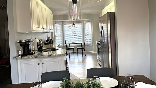 A modern kitchen with white cabinets, a dining area with a table set for four, and a large window with blinds. - King Bed, Workspace, Games near RDU, RTP, Downtown (Raleigh)