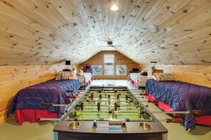 Une pièce mansardée chaleureuse avec des murs en bois et un plafond en pente. Elle comprend deux lits avec une literie rouge et bleue, une table de baby-foot et une fenêtre donnant sur les arbres.