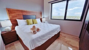 Una cama bien hecha con una colcha blanca y dos toallas enrolladas, un cabecero de madera, una mesita de noche con una lámpara, y una ventana con vistas al paisaje.