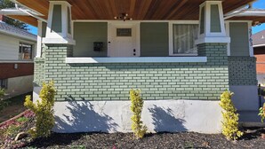 A single-story house with a green brick facade, a white door, and a covered porch with a wooden roof. - Charming Bungalow • Hot Tub • Near Trails & Dining (Ogden)