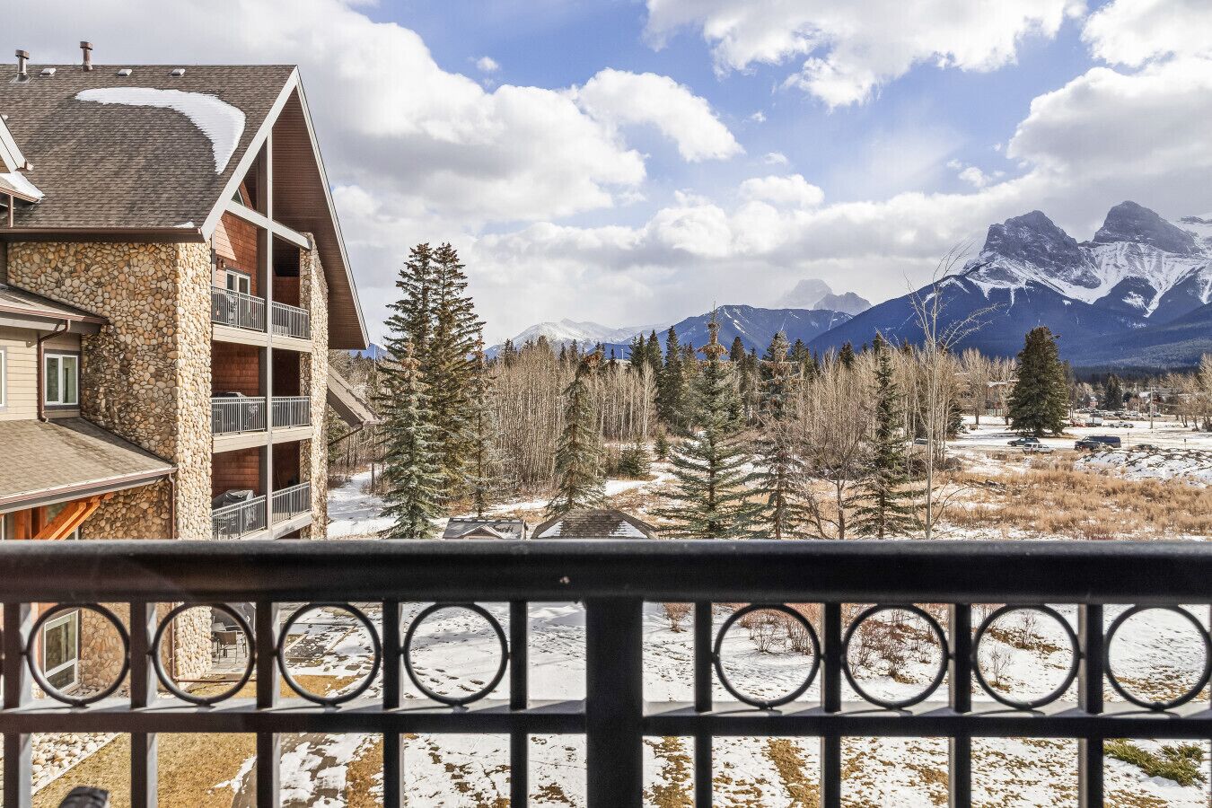 A mountain lodge with a stone facade, a balcony with metal railings, and a view of snow-covered mountains.