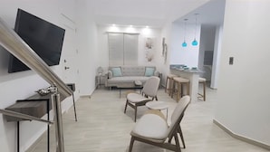Ein modernes Wohnzimmer mit einer Couch, Sesseln, einem Couchtisch, einem an der Wand montierten Fernseher und einer Küchenzeile mit Barhockern und einer Arbeitsfläche.