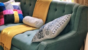 A teal sofa with a yellow throw blanket and patterned pillows, positioned in front of a window with a view of greenery.
