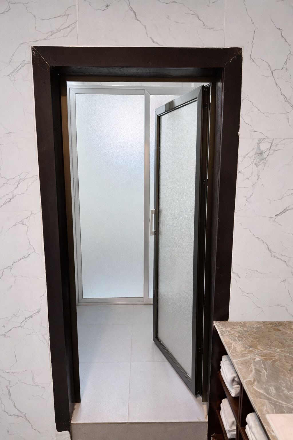Un couloir avec une porte en verre dépoli, un mur en marbre et une étagère en bois où sont posées des serviettes.