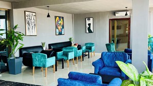 Un moderno vestíbulo de hotel con un sofá de cuero negro, sillones azul turquesa y azul, un mostrador de recepción de mármol y varias plantas en macetas.