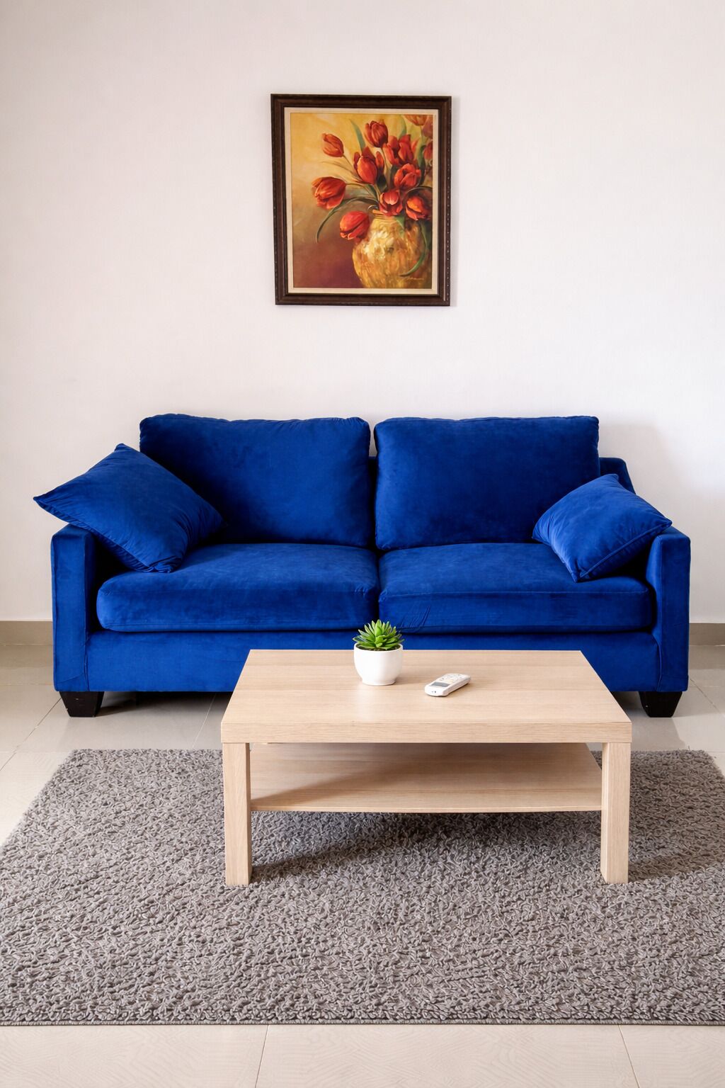 Een blauwe bank met daarboven een ingelijst schilderij, een klein tafeltje met een plant en een afstandsbediening, en een grijze vloerkleed.