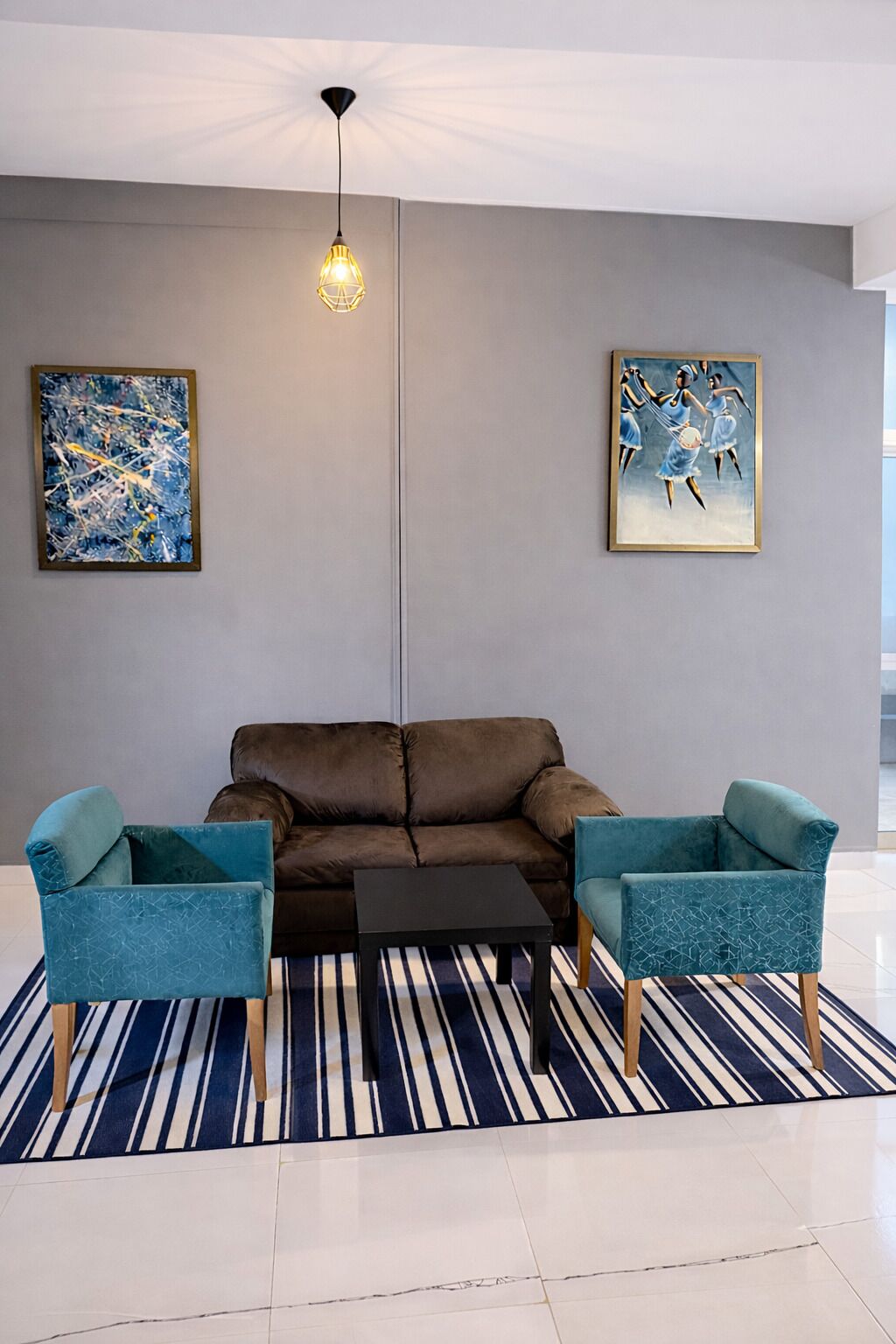 A modern living room with a brown sofa, two teal armchairs, a black coffee table, and a striped rug. There are two framed artworks on the walls.