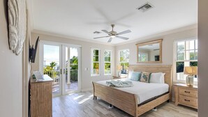 Ein Schlafzimmer mit einem hölzernen Bett, einem Deckenventilator, einem großen Fenster und einem Balkon mit Blick auf Grünflächen.