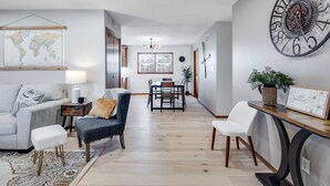 Un salon moderne comprenant un canapé, un fauteuil et un pouf, un coin repas avec une table et des chaises, et une grande horloge murale.