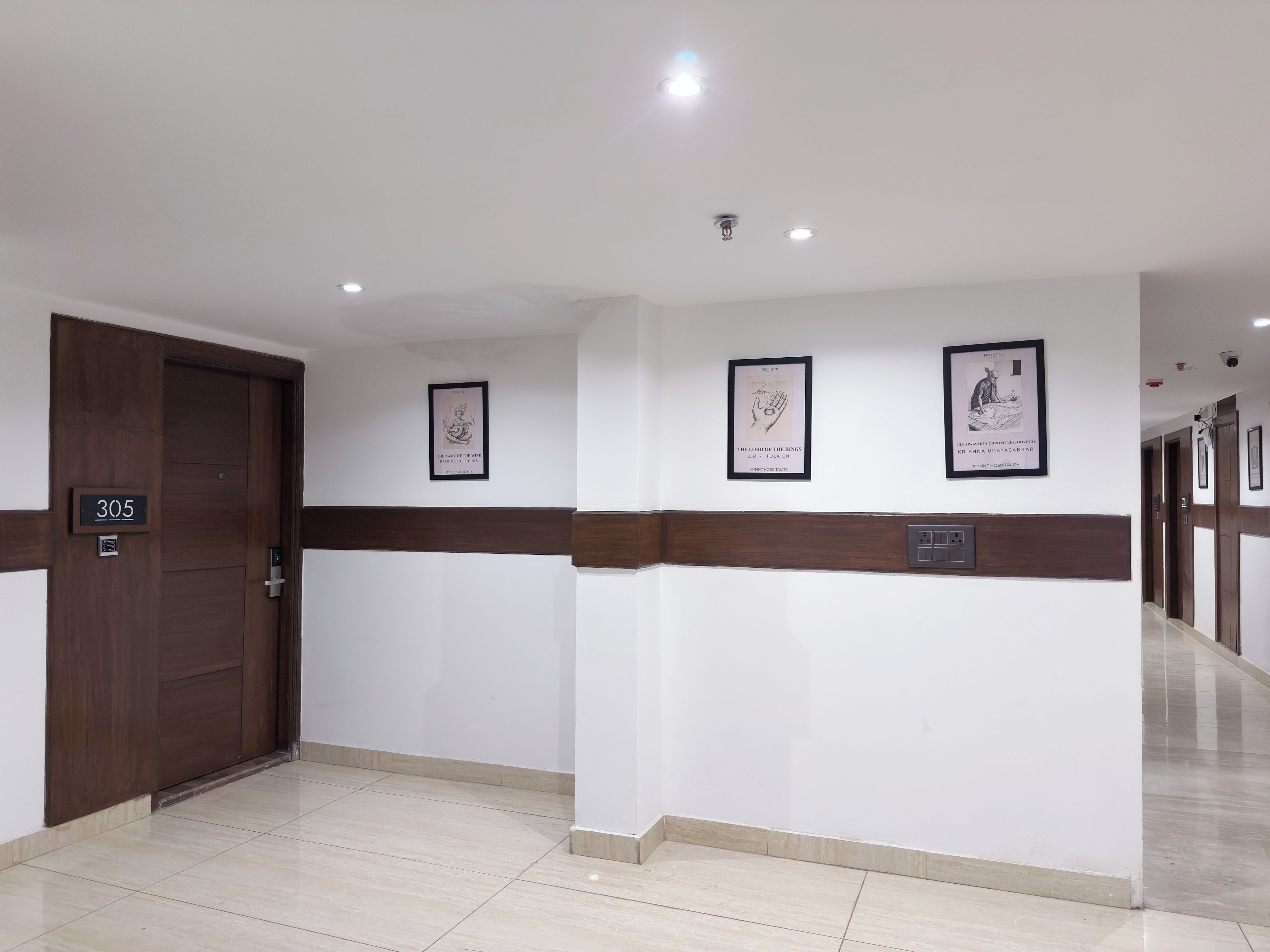 A hotel lobby with a wooden door marked '305', a tiled floor, and three framed pictures on the wall.