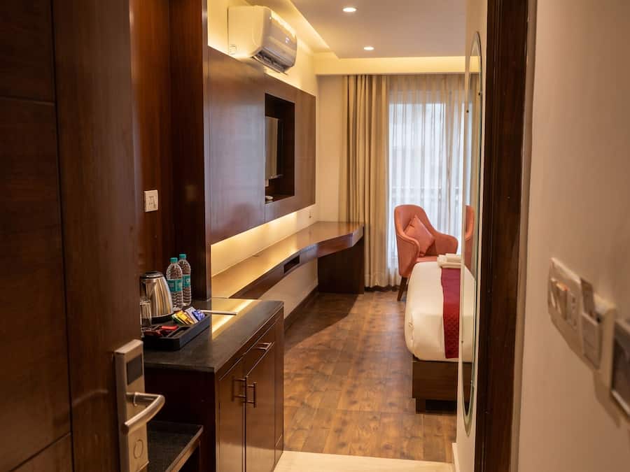 A hotel room with a wooden desk, a flat-screen TV mounted on the wall, a bed with a red blanket, and a window with curtains.