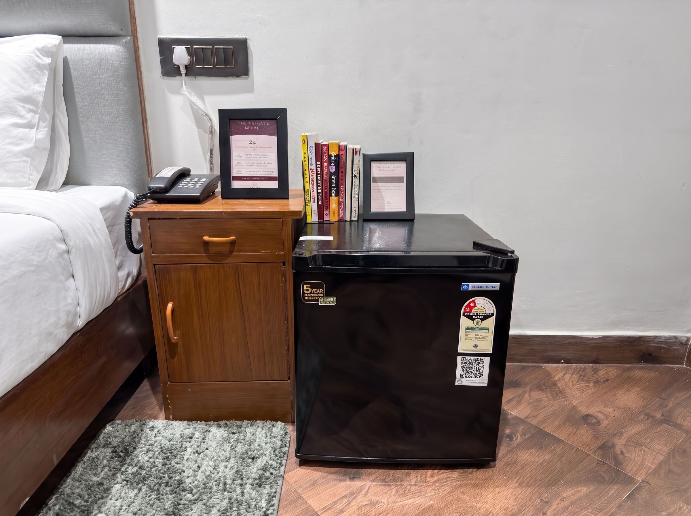 Een klein houten kastje met een lade, een zwarte koelkast met stickers, een ingelijst certificaat en een telefoon op een nachtkastje.