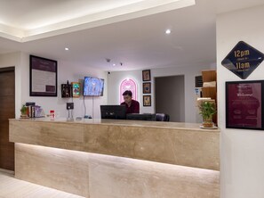 Zona de recepción de un hotel con mostrador de mármol, pantalla digital y letrero de neón.