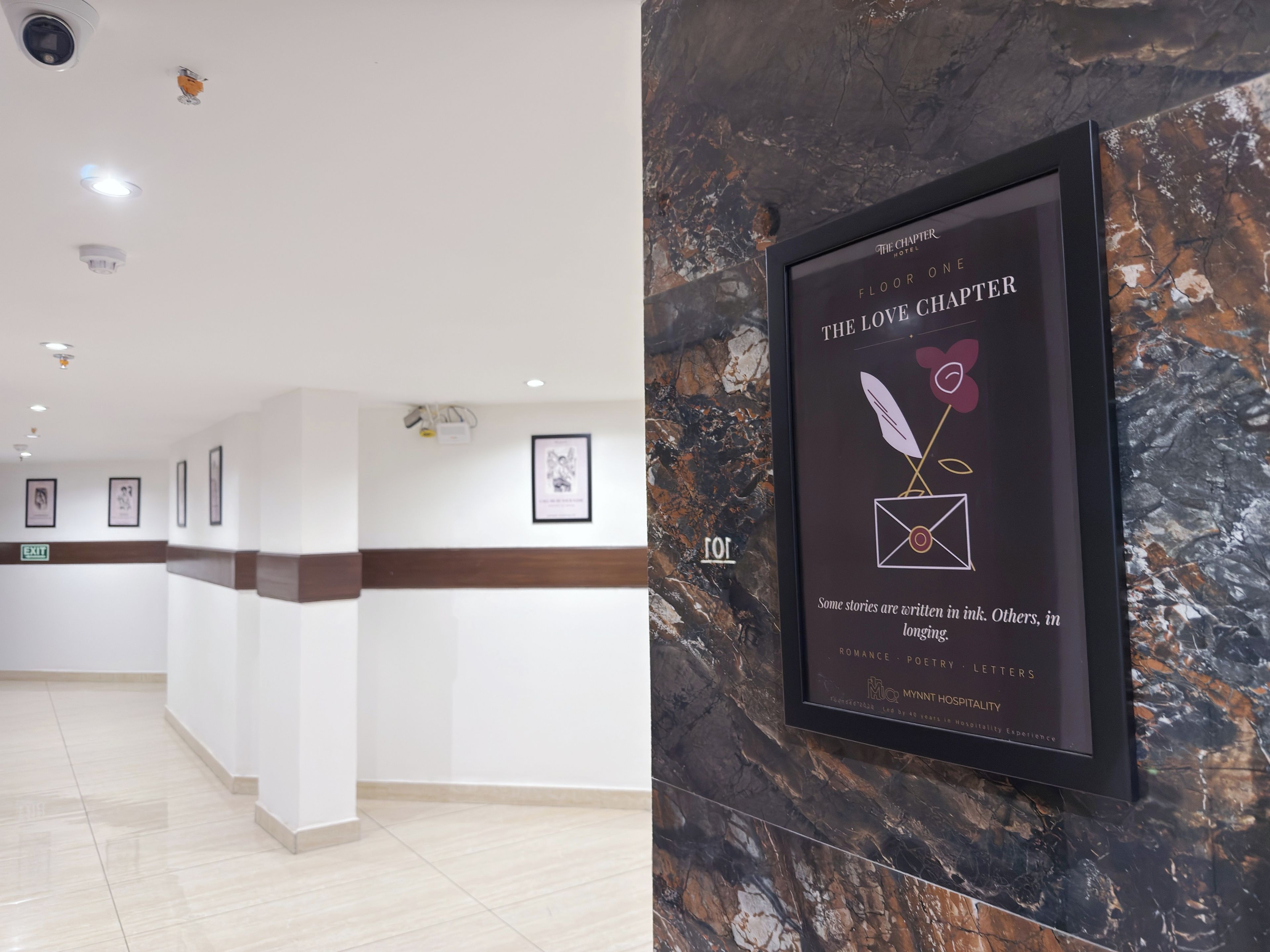 Uma área de recepção de hotel com parede de mármore, obras de arte emolduradas e uma placa com o nome e a localização do hotel.