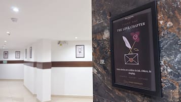Uma área de recepção de hotel com parede de mármore, obras de arte emolduradas e uma placa com o nome e a localização do hotel.