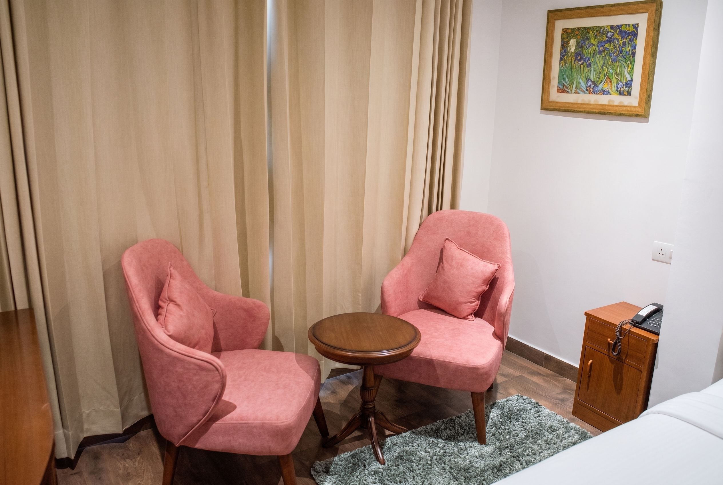 A room with two pink armchairs, a small wooden table, a wooden nightstand with a phone, and a painting on the wall.