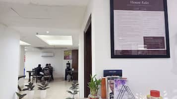 Zona de recepción de hotel con un documento de normas internas enmarcado, mostrador de recepción, plantas en macetas y un área de descanso al fondo.