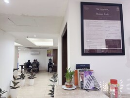 A hotel reception area with a framed house rules document, a reception desk, potted plants, and a seating area in the background.
