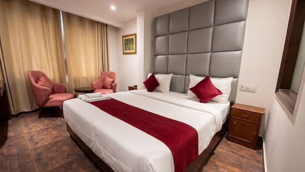 Hotellihuone, jossa on suuri sänky, kaksi vaaleanpunaista nojatuolia, pieni pöytä ja pääty, jossa on kuvioitu, tikattu kangas.