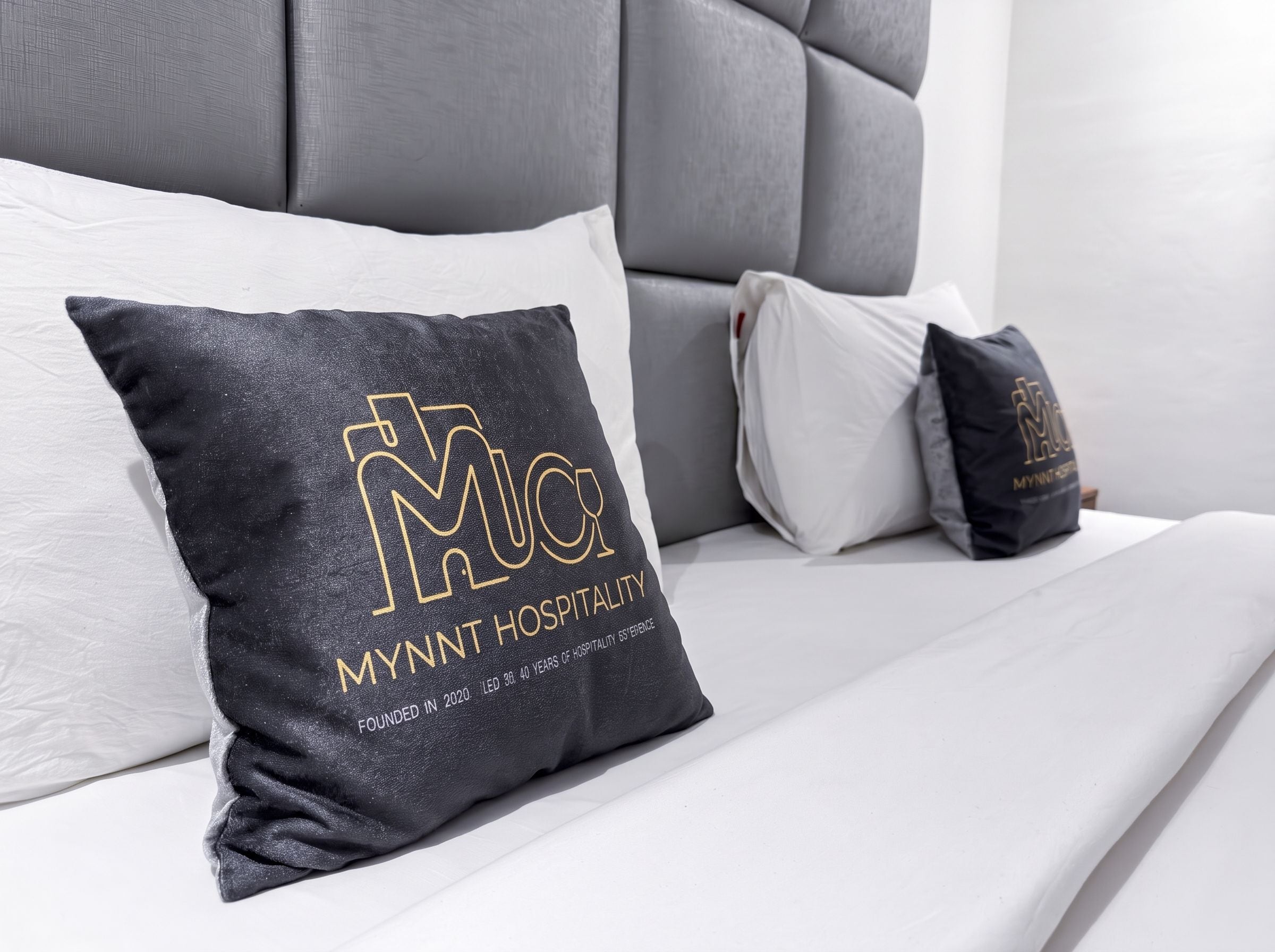 A bed with a grey headboard and multiple pillows, including a black pillow with gold lettering and text.