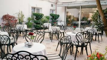 An outdoor dining area with round tables and chairs, surrounded by potted plants and a patterned floor.
