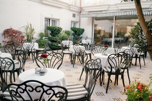 An outdoor dining area with round tables and chairs, surrounded by potted plants and a patterned floor.