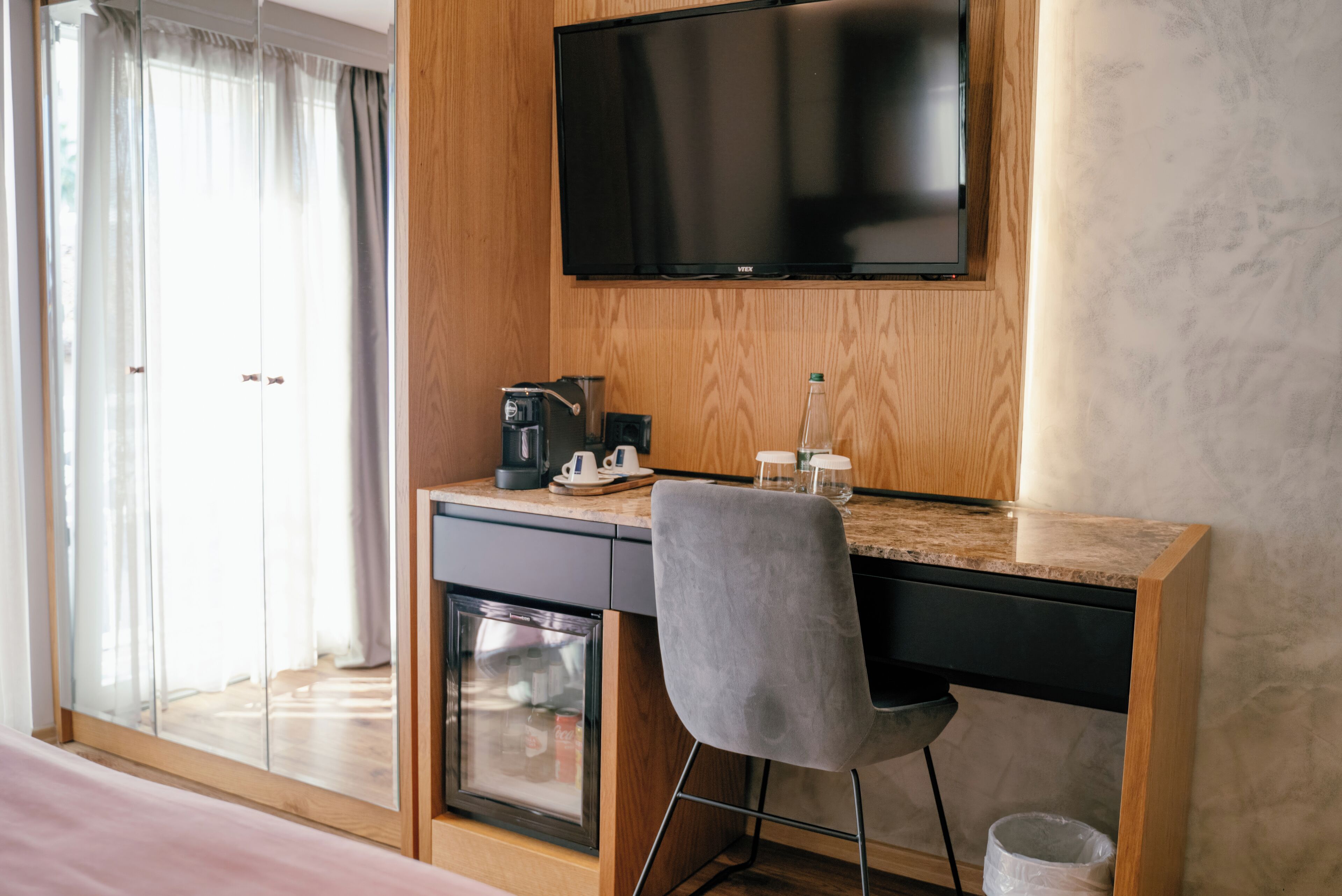Habitación de hotel con escritorio de madera, televisión montada en la pared, cafetera y un refrigerador pequeño.