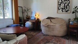 Una sala de estar acogedora con una mesa redonda para café, un sillón orejero color beige, un escritorio con una computadora portátil y un cuadro abstracto de gran tamaño en la pared.