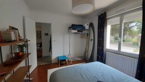 Un dormitorio con cama, una estantería con libros y una planta, una ventana con cortinas y una puerta que da a otra habitación.
