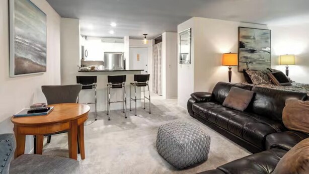 Ein modernes Wohnzimmer mit Ledersofa, rundem Couchtisch und grauem Hocker. Im Hintergrund ist eine Küchenzeile mit Bar und Hockern erkennbar.