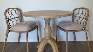 Una mesa de comedor con dos sillas de mimbre, cada una con un cojín, frente a una pared lisa con tres cuadros de paisajes desérticos enmarcados encima.