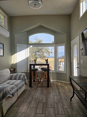 A room with a wooden floor, a sofa, a table with stools, and a view through large windows. - Rural Lincoln Tiny House - country feel with fast freeway access (Lincoln)