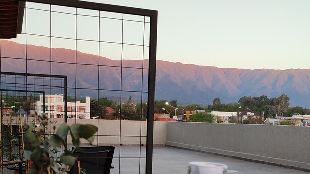 Une table en verre sur laquelle se trouvent un pot de plante, un verre d’eau, un verre contenant une boisson rouge et une canette de soda rouge. En arrière-plan, on aperçoit des montagnes et un bâtiment.