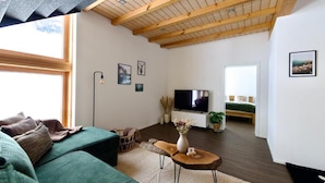 En moderne stue med en grøn sofa, træloft, et fladskærms-tv og en seng i baggrunden.