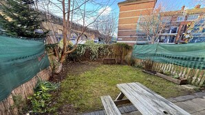 A backyard with a wooden picnic table, a grassy area, and a brick building in the background. - Central London Apartment Near Tower Bridge | Private Entrance & Garden (London)
