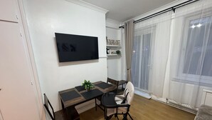 A modern living room with a wooden dining table, a flat-screen TV mounted on the wall, and a large window with sheer curtains. - Central London Apartment Near Tower Bridge | Private Entrance & Garden (London)
