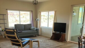 A living room with a sofa, a TV, a wooden chair, and a large window. - Sunny & Private I bedroom House with Sun Room for Guests (Torrey)