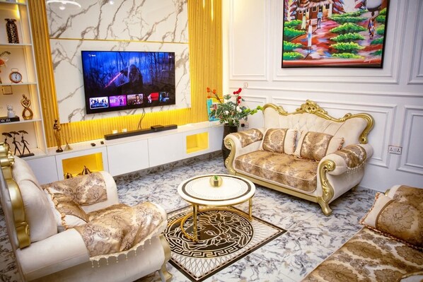 A luxurious living room with a marble floor, a gold-framed sofa, a white armchair, a round coffee table, and a flat-screen TV mounted on the wall.
